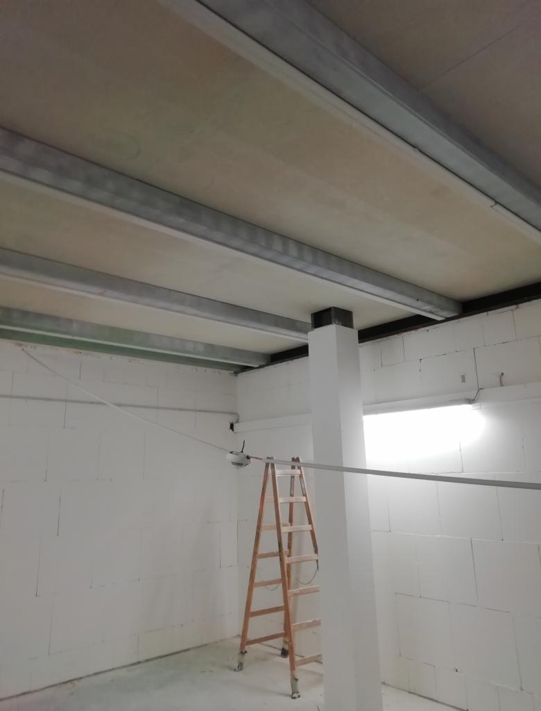 Montaż szkieletu sufitu podwieszanego z metalowych profili i płyt w pomieszczeniu z białymi ścianami z betonu komórkowego, widoczna drabina oparta o filar.