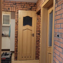 Jacek Maciołek - Wnętrze mieszkania z ceglaną ścianą, drewnianymi drzwiami z szybą i widoczną kuchnią w tle; na podłodze skrzynka z narzędziami.