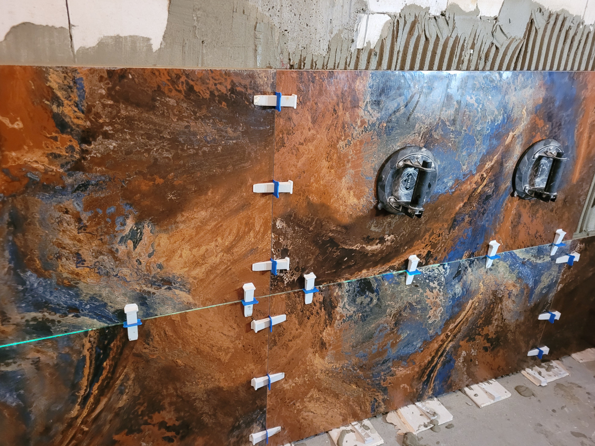 Układanie wielkoformatowych płytek imitujących rdzę i miedź, z użyciem przyssawek i systemu poziomowania z niebieskimi klinami, widoczny laserowy wskaźnik poziomu i surowa ściana w tle.