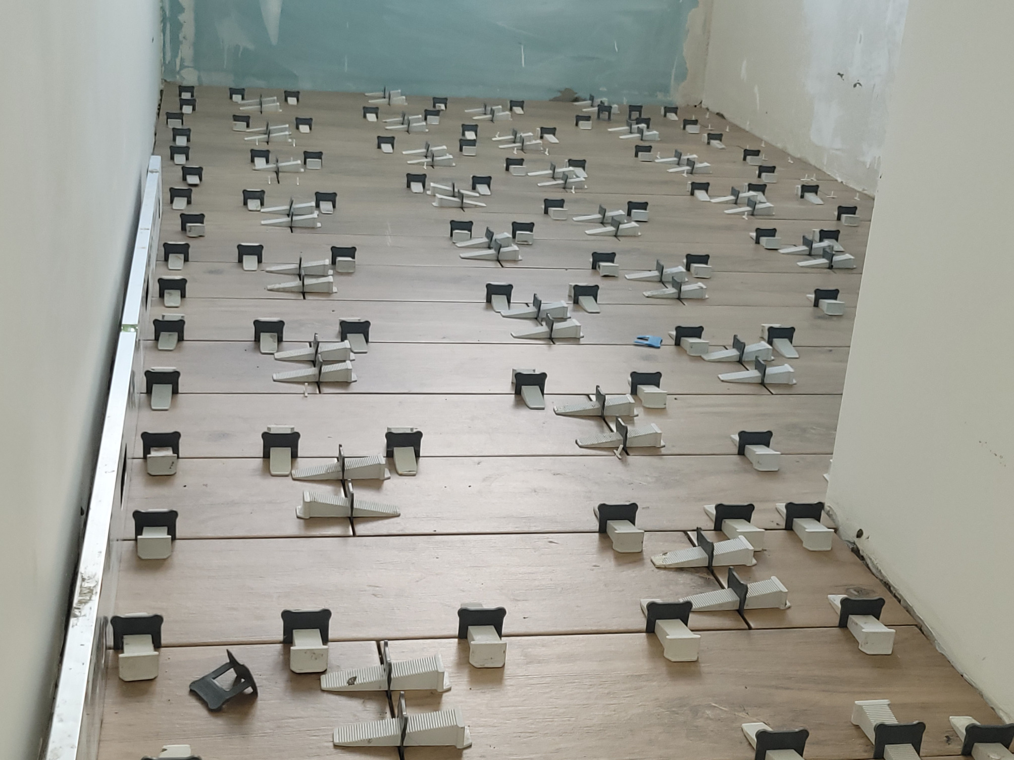 Układanie płytek podłogowych z użyciem systemu poziomowania, widoczne klipsy i kliny, ściany w trakcie przygotowania.