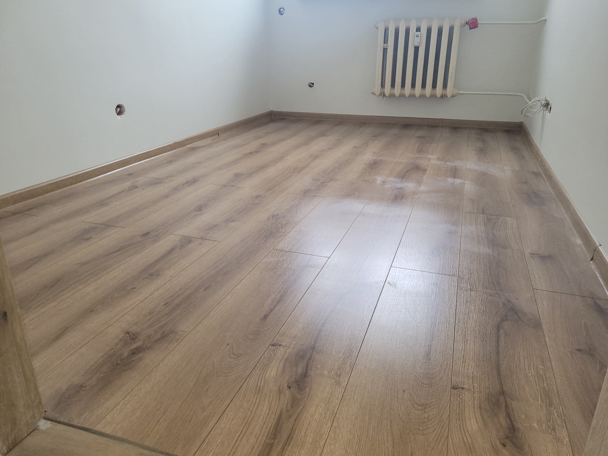 Pokój po remoncie z nową, jasnobrązową podłogą laminowaną, białe ściany z widocznymi puszkami elektrycznymi bez osprzętu i stary, żeliwny grzejnik.