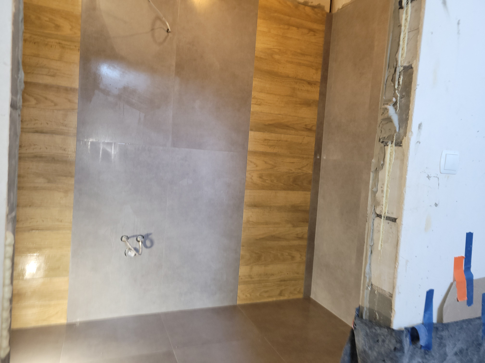 Wykończona łazienka z mieszanką płytek imitujących drewno i szary beton. Widoczny montaż baterii prysznicowej i fragment niezabezpieczonej ściany z instalacją.