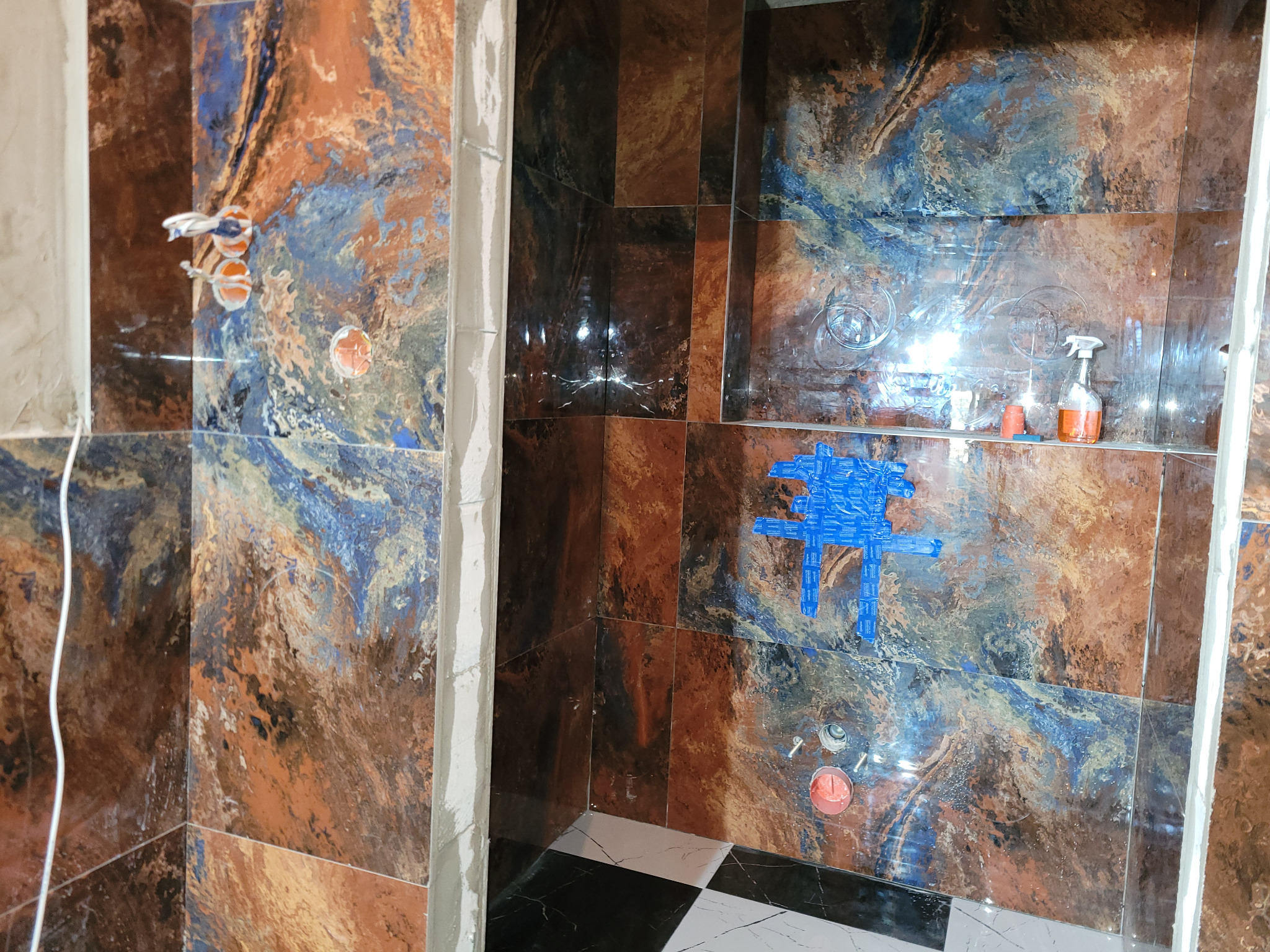 Montaż płytek w łazience z wzorem marmuru w odcieniach brązu i niebieskiego. Widoczne instalacje wodne i fragment podłogi z czarno-białej szachownicy.