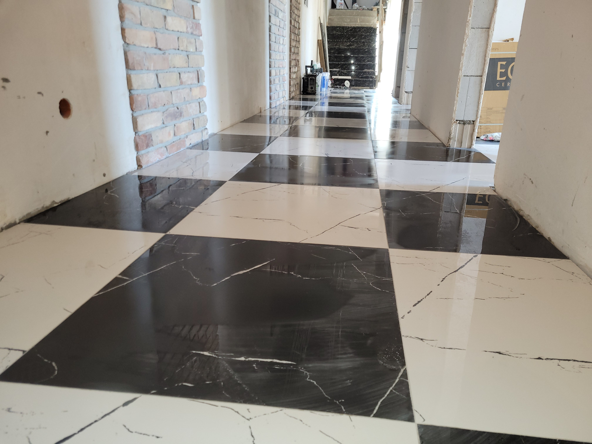 Błyszcząca, czarno-biała podłoga w szachownicę, wykończona w korytarzu z fragmentami ściany z cegły i surowego tynku. Widoczne puste przestrzenie po instalacjach.