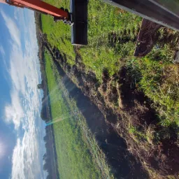 Wykopany rów w ziemi, widoczny fragment koparki z łyżką, tło stanowi zielona łąka i błękitne niebo z chmurami, robota ziemna w toku.