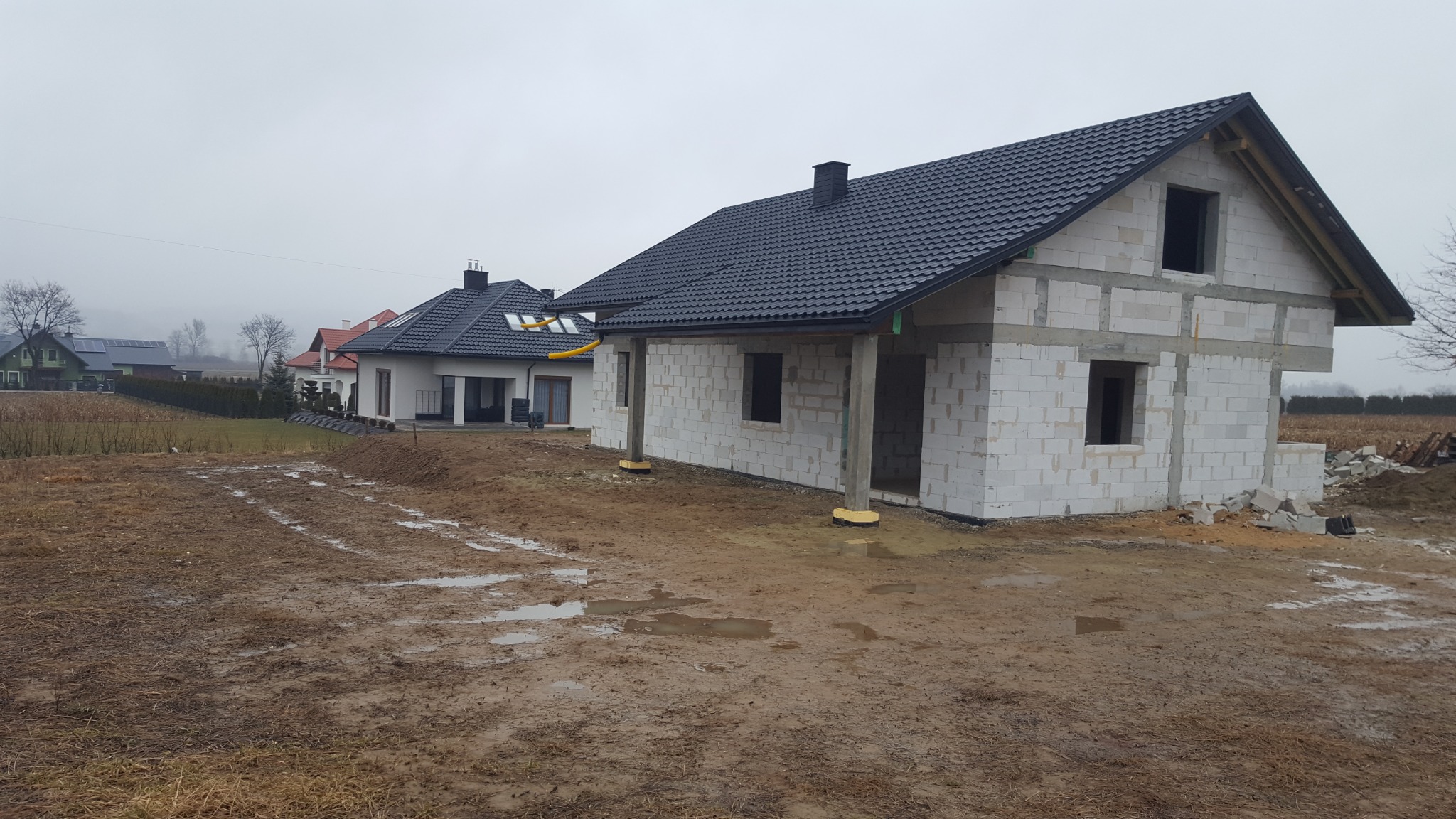 Dom murowany w stanie surowym zamkniętym z czarną dachówką, widoczne bloczki betonowe i otwory okienne, na pierwszym planie błotniste podłoże, w tle inne domy.