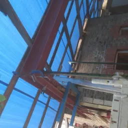 Wnętrze budynku w trakcie budowy więźby dachowej, widoczne stalowe elementy konstrukcyjne, rusztowania i niebieska plandeka ochronna.