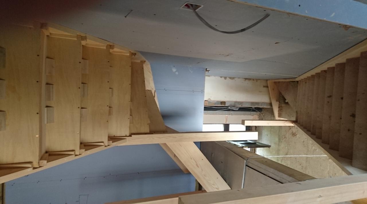 Wnętrze w trakcie budowy z widoczną drewnianą konstrukcją schodów, surowym sufitem z wystającym kablem i niebieską ścianą z płyt gipsowo-kartonowych.