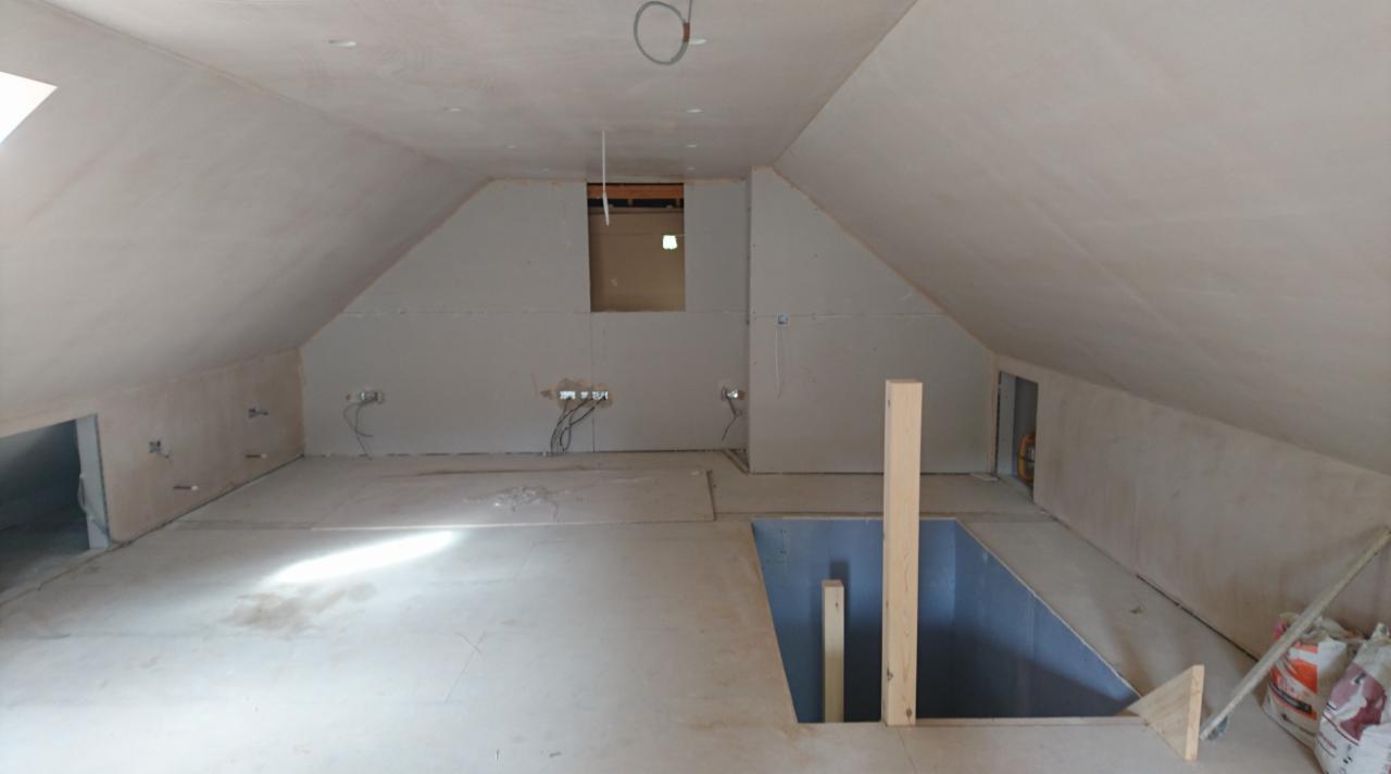 Surowe poddasze w trakcie adaptacji w Gliwicach. Widoczne otynkowane skosy dachu, przygotowane otwory na instalacje elektryczne, częściowo położona podłoga oraz otwór w podłodze zabezpieczony...