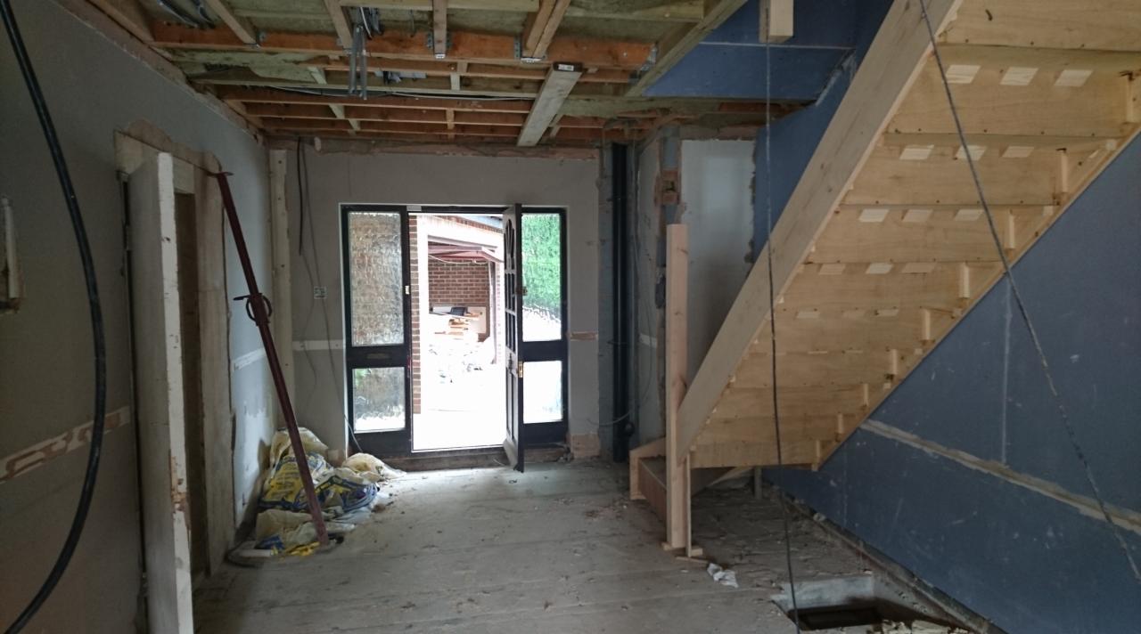 Wnętrze w trakcie remontu: widoczne surowe ściany, drewniana konstrukcja schodów, odsłonięty strop z instalacjami i otwarte drzwi wejściowe z widokiem na podwórze.