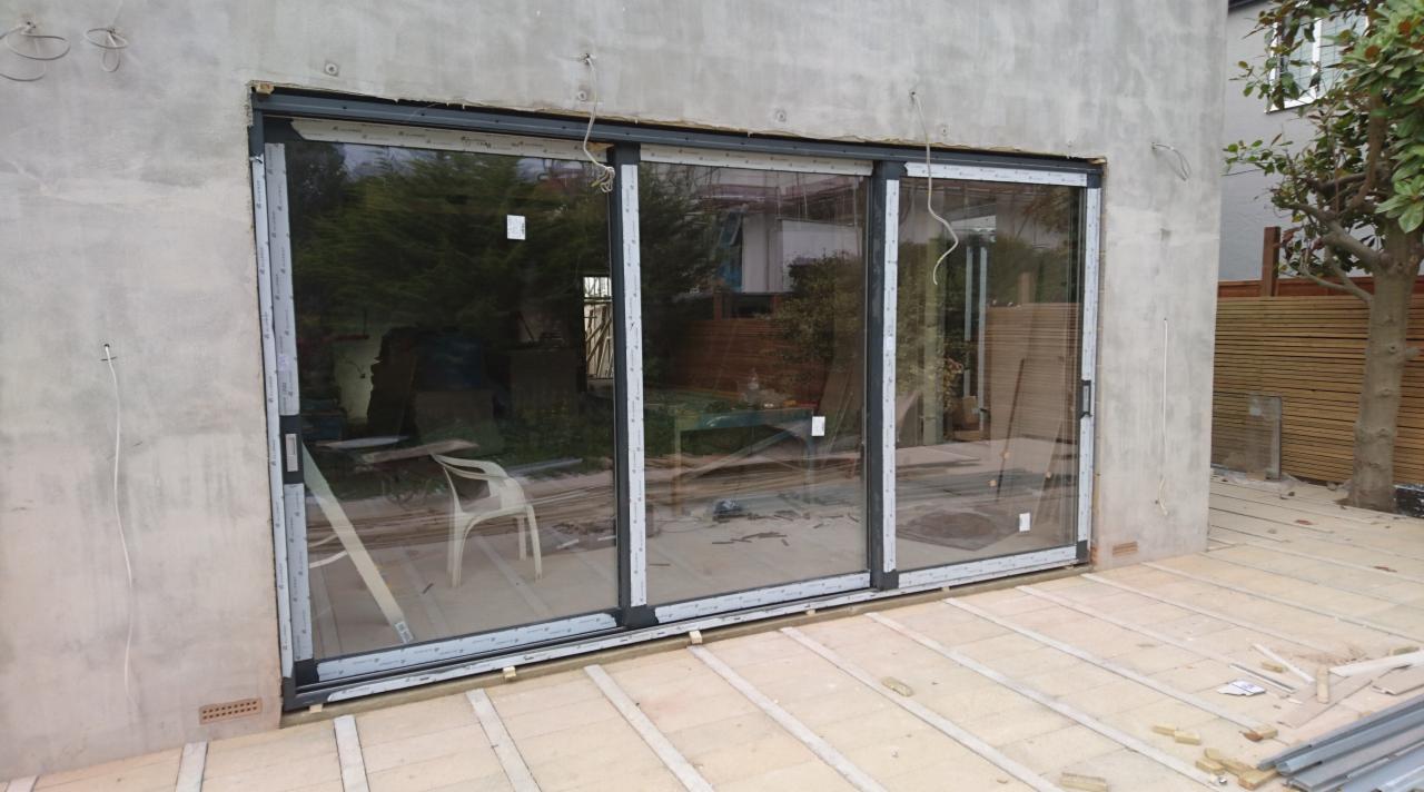 Montaż trzyskrzydłowych, przesuwnych drzwi tarasowych z aluminiową ramą w surowym stanie, widoczne niedokończone podłoże tarasu i otynkowana ściana budynku z wystającymi przewodami.