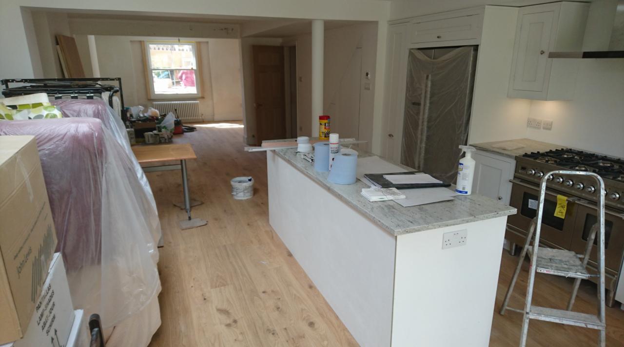 Wnętrze kuchni w trakcie remontu: meble zabezpieczone folią, wyspa kuchenna z blatem z jasnego granitu, kuchenka gazowa z drabinką, podłoga z paneli. Widoczny bałagan remontowy.