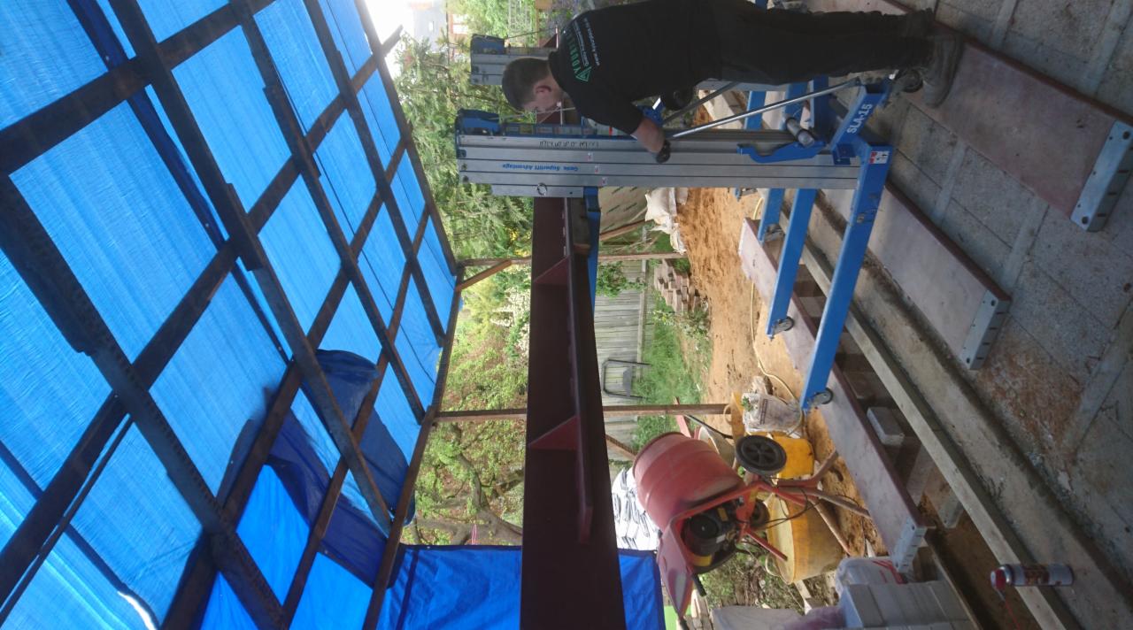 Pracownik montuje element konstrukcyjny dachu z użyciem podnośnika, widoczna więźba dachowa zabezpieczona niebieską plandeką, w tle betonowa podstawa i mieszalnik.