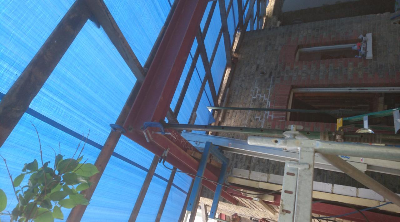 Wnętrze budynku w trakcie budowy więźby dachowej, widoczne stalowe elementy konstrukcyjne, rusztowania i niebieska plandeka ochronna.