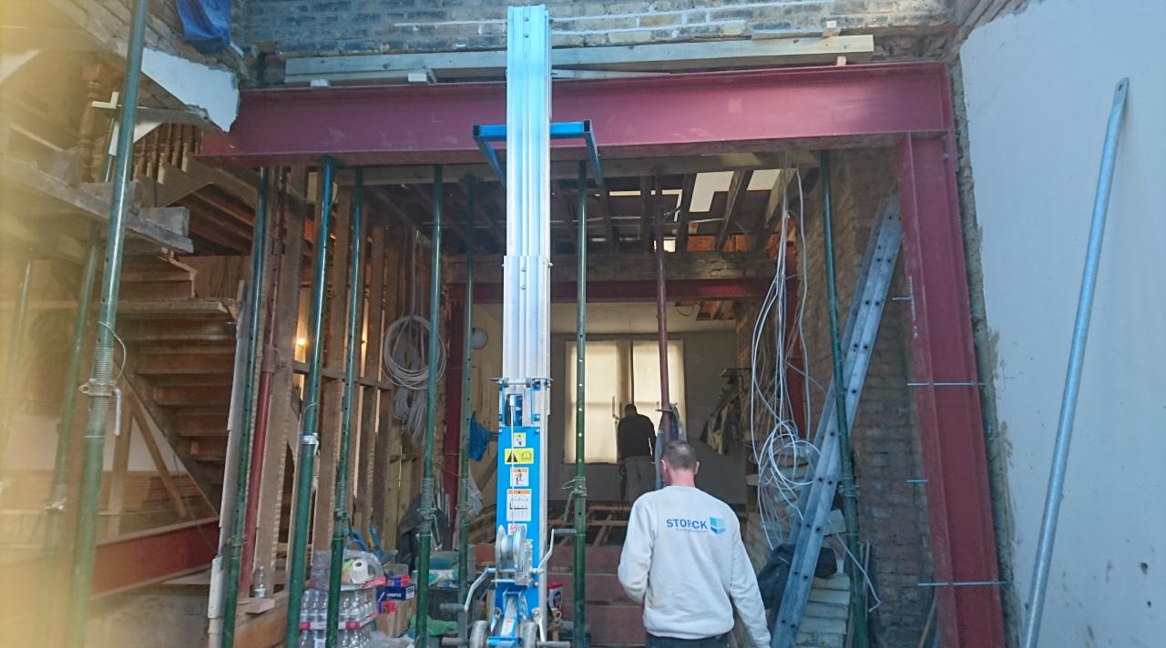 Wnętrze w trakcie remontu z odsłoniętą cegłą, stalową belką nośną podpartą stemplami, podnośnikiem platformowym i fragmentem drewnianych schodów.