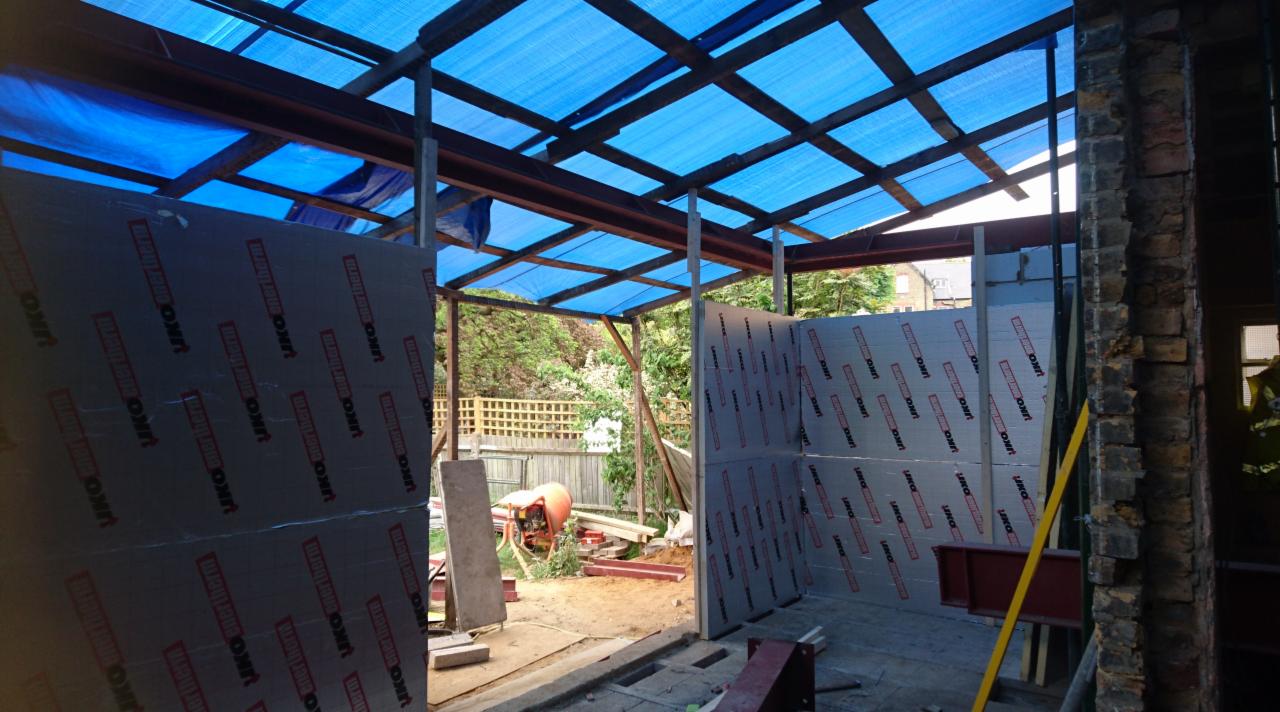 Budowa zadaszenia z blachy falistej na metalowej konstrukcji, ściany ocieplone płytami izolacyjnymi z widocznym logo producenta, ceglana ściana w tle, betoniarka i materiały budowlane na zewnątrz.