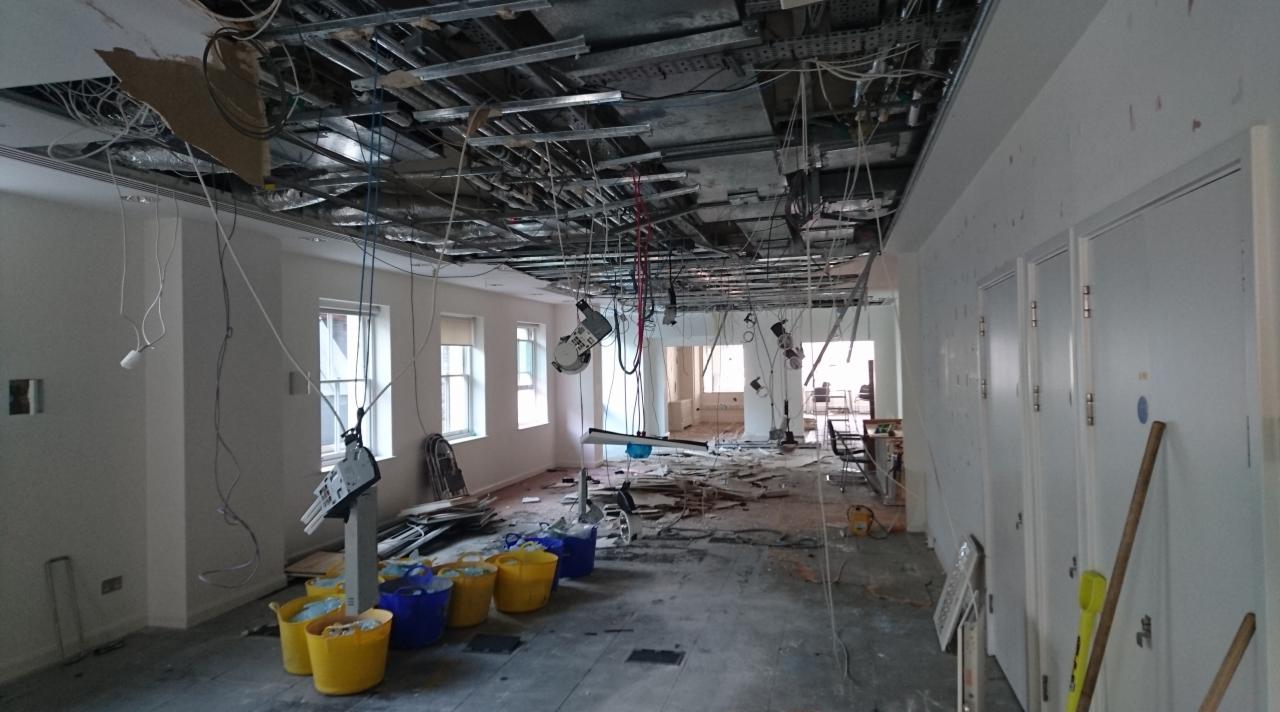 Wnętrze biura w trakcie demontażu: odsłonięty sufit z wiszącymi przewodami, gruz na podłodze i wiadra na odpady.