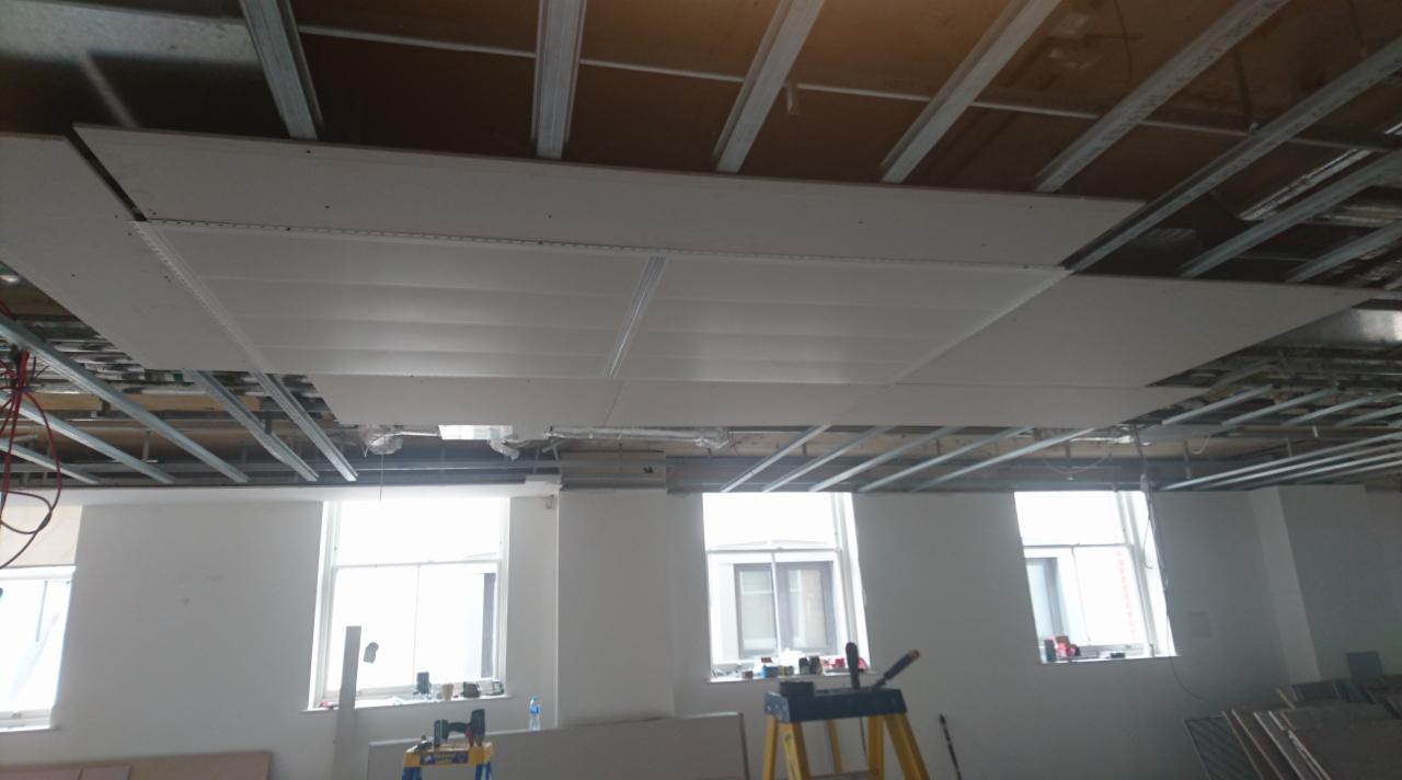Montaż sufitu podwieszanego z płyt gipsowo-kartonowych na metalowej konstrukcji w pomieszczeniu z oknami, widoczne narzędzia i materiały budowlane.