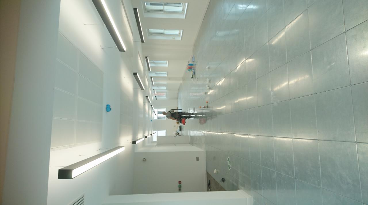 Wnętrze biura w trakcie wykańczania: pracownik na podłodze, widoczne panele ścienne, sufit z oświetleniem liniowym i okna.