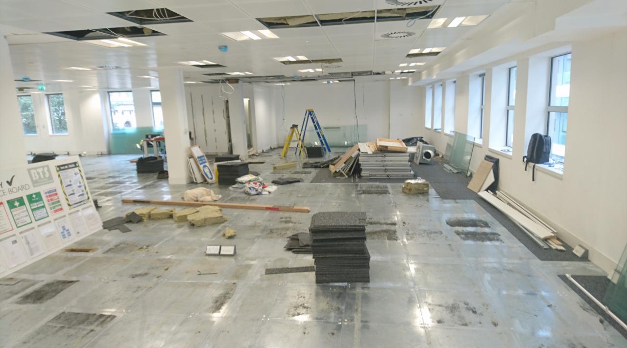 Wnętrze biura w trakcie remontu, widoczne rozłożone panele podłogowe, drabiny, materiały budowlane i odsłonięty sufit podwieszany.
