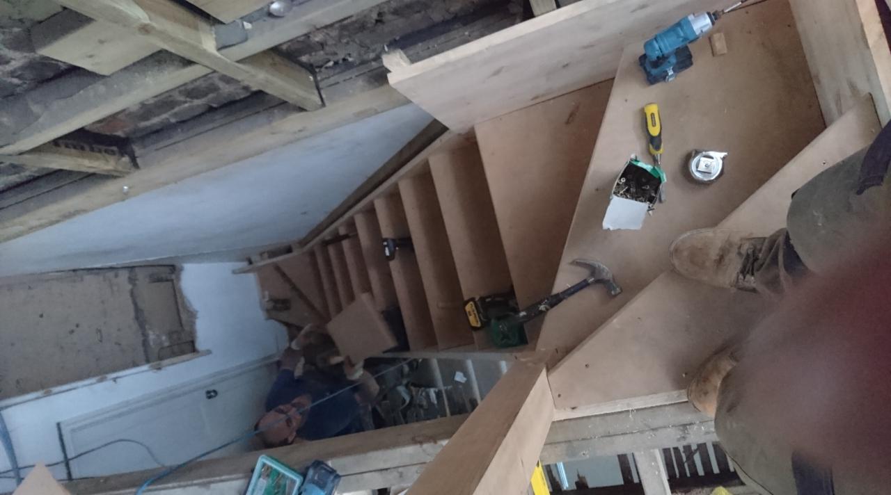 Montaż drewnianych schodów w surowym stanie, widoczne narzędzia: wiertarka, młotek, miarka oraz instalacja elektryczna w puszce, widok z góry na pracownika w trakcie pracy.