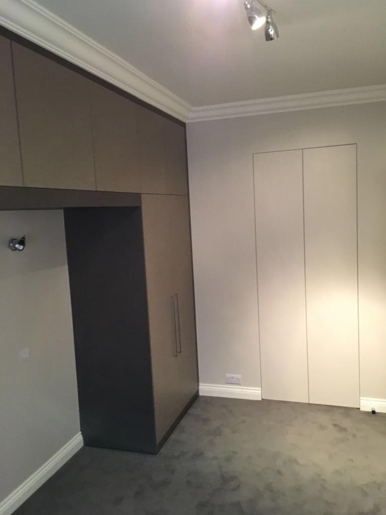 Wnętrze pokoju z zabudowaną szafą w odcieniach szarości i beżu, szary dywan na podłodze, białe listwy przypodłogowe i ozdobne listwy przysufitowe, oświetlenie punktowe na suficie.