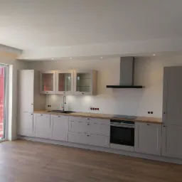 Jasna kuchnia z drewnianym blatem, czarną armaturą i szklanymi witrynami w górnych szafkach, widok na balkon przez duże okno.