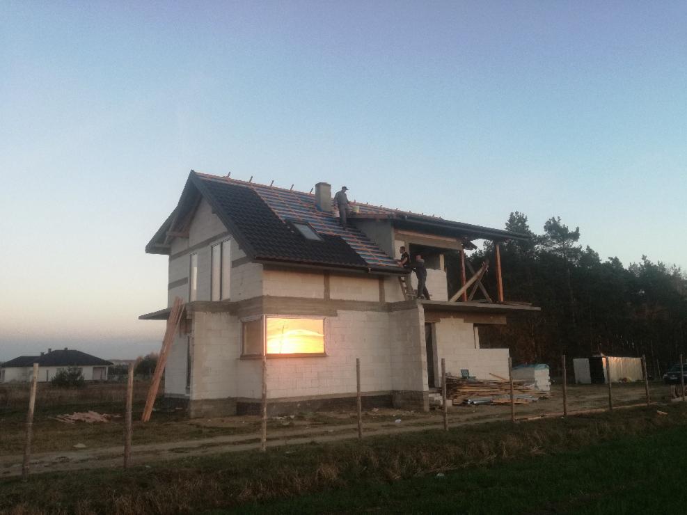 Budowa domu jednorodzinnego w trakcie realizacji: dekarze układają dachówkę na częściowo ukończonym dachu, widoczne bloczki betonowe ścian i drewniane elementy konstrukcyjne, w oknie odbija się...