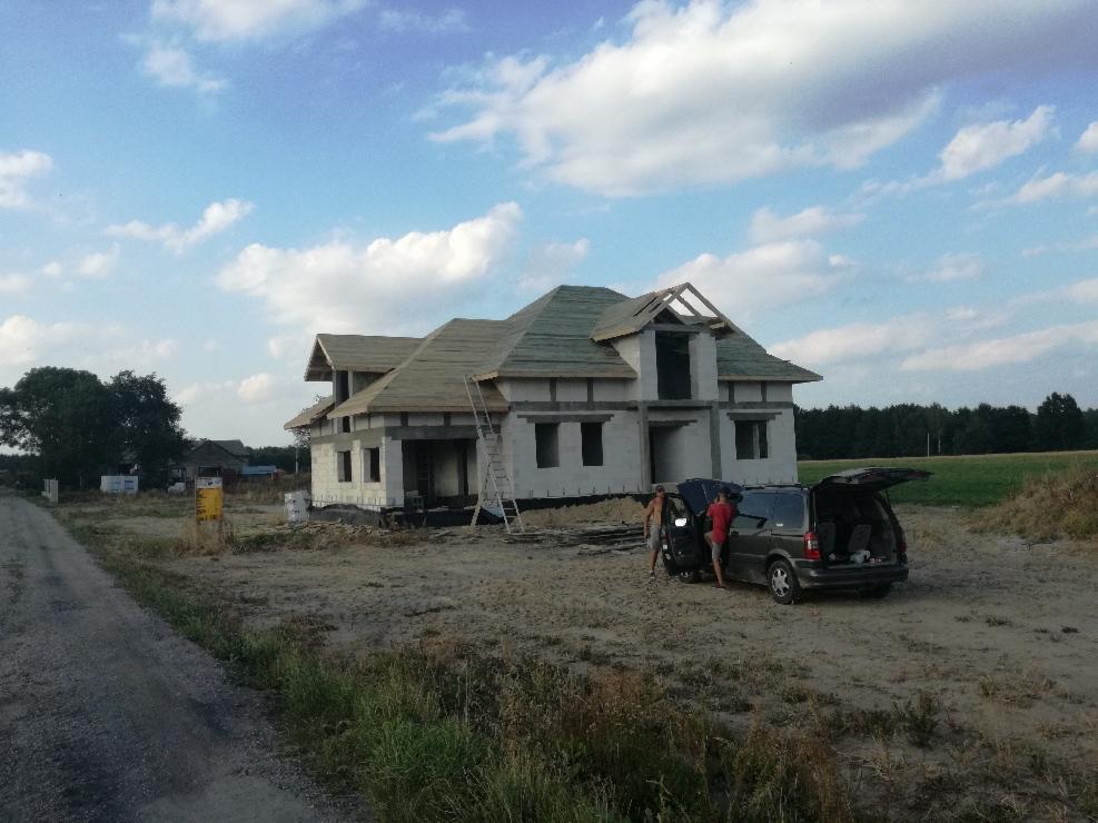 Surowy stan dwukondygnacyjnego domu z częściowo położonym dachem z desek, drabina oparta o ścianę, obok zaparkowany van z otwartym bagażnikiem i dwoma osobami.