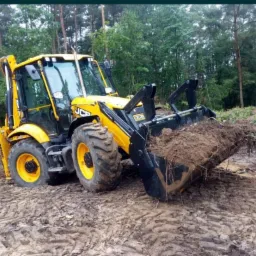 Żółto-czarna koparko-ładowarka JCB z pełną łyżką ziemi na błotnistym terenie, w tle zielony las.