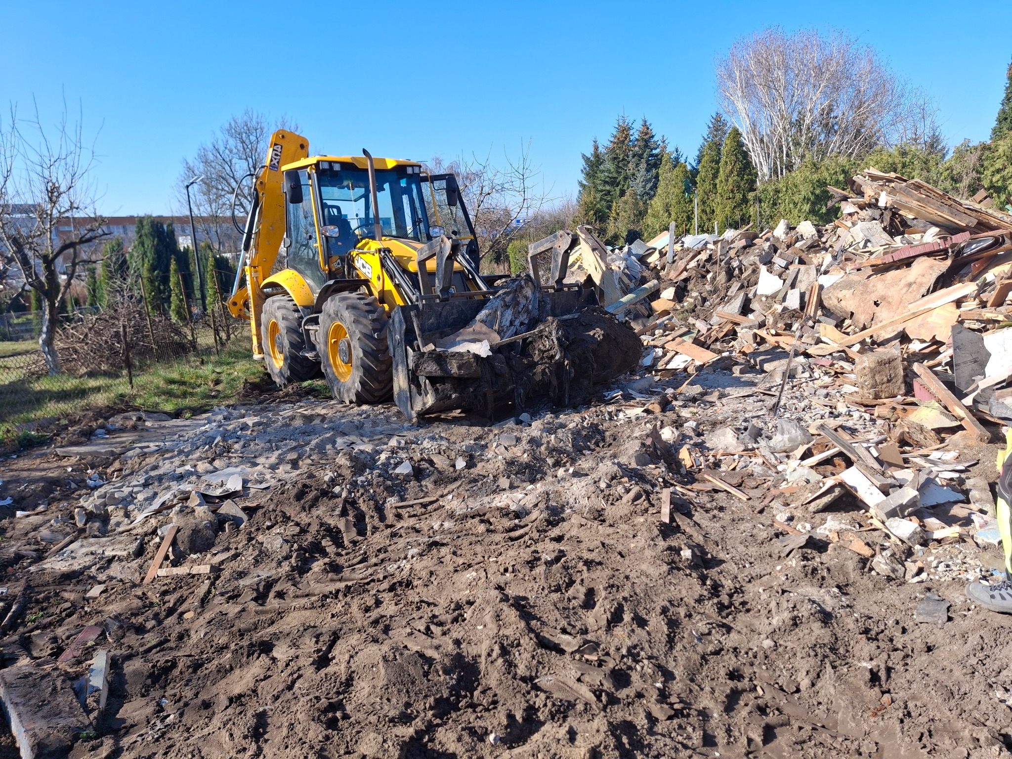 Żółta koparko-ładowarka JCB z łyżką pełną gruzu i ziemi na terenie po rozbiórce, w tle sterta pozostałości po budynku, drzewa i błękitne niebo.