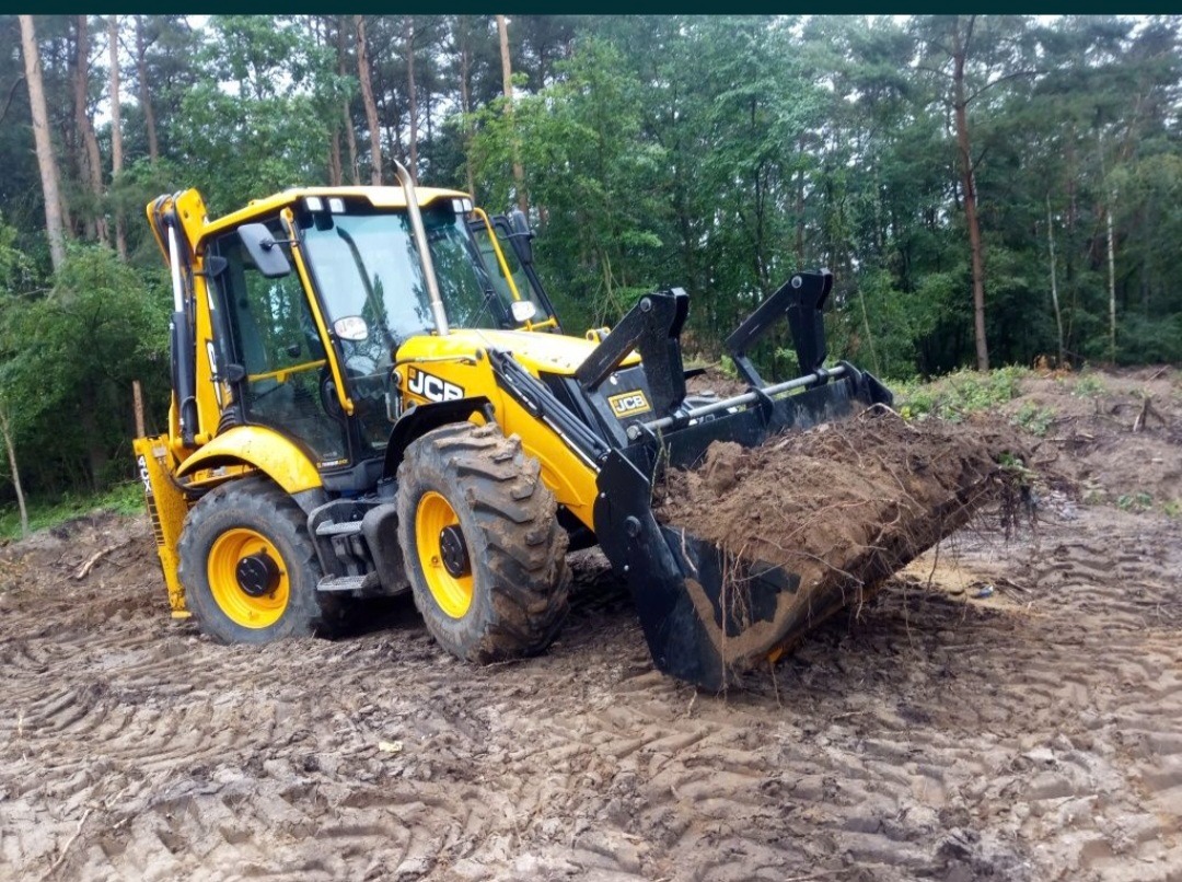 Żółto-czarna koparko-ładowarka JCB z pełną łyżką ziemi na błotnistym terenie, w tle zielony las.