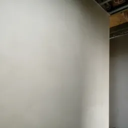 Świeżo położone, jasne, gładkie wykończenie ściany w trakcie remontu, widoczne surowe elementy konstrukcyjne sufitu.