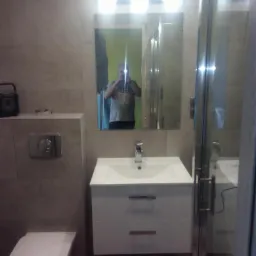 Nowoczesna łazienka z umywalką w szafce z dwoma szufladami, wiszącą toaletą i kabiną prysznicową; widoczne szare płytki na ścianach i drewnopodobna podłoga; odbicie fotografa w lustrze.