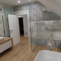 Nowoczesna łazienka z drewnianą podłogą, szklaną kabiną prysznicową i umywalką na szafce z drewnianym blatem. W tle widoczny grzejnik drabinkowy i otwarte drzwi do innego pomieszczenia.