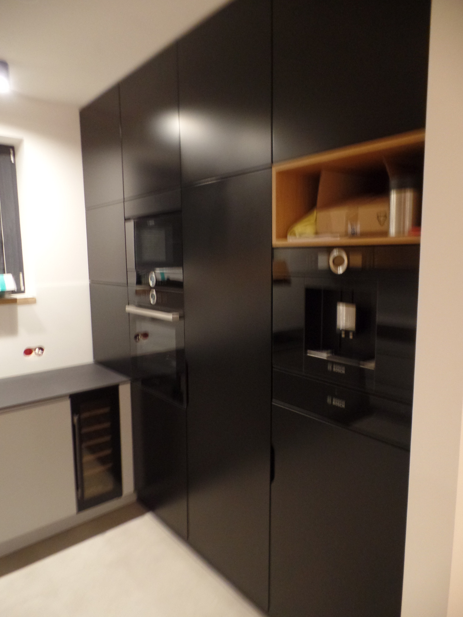 Część zabudowy kuchennej z czarnymi frontami, zawierająca wbudowany piekarnik, ekspres do kawy i szafkę na wino, z widocznymi akcesoriami kuchennymi w otwartej półce.