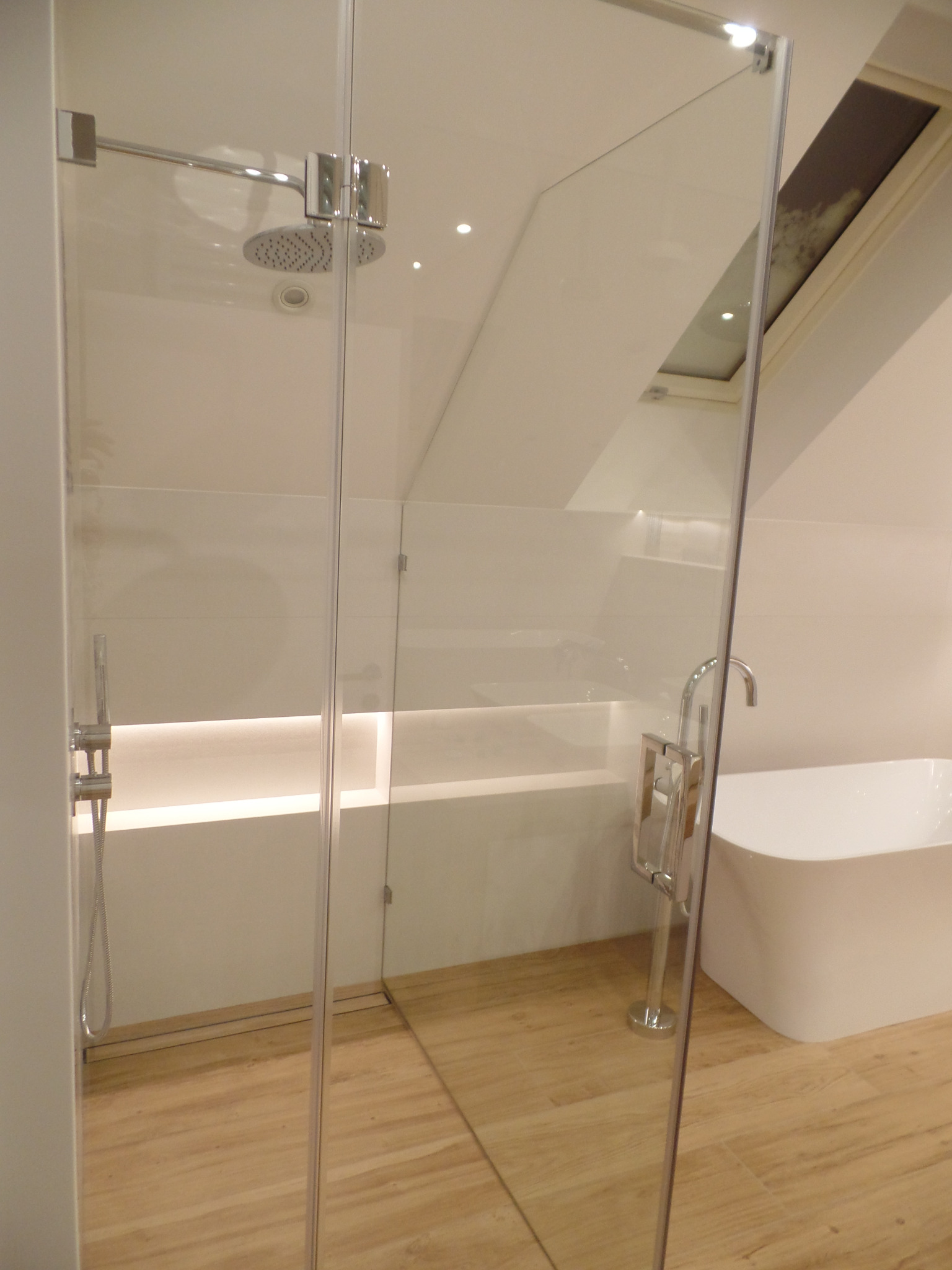 Nowoczesna kabina prysznicowa z deszczownicą i chromowanymi okuciami, widoczna wanna wolnostojąca i oświetlenie LED wnęki, podłoga wyłożona płytkami imitującymi drewno, okno dachowe w tle.