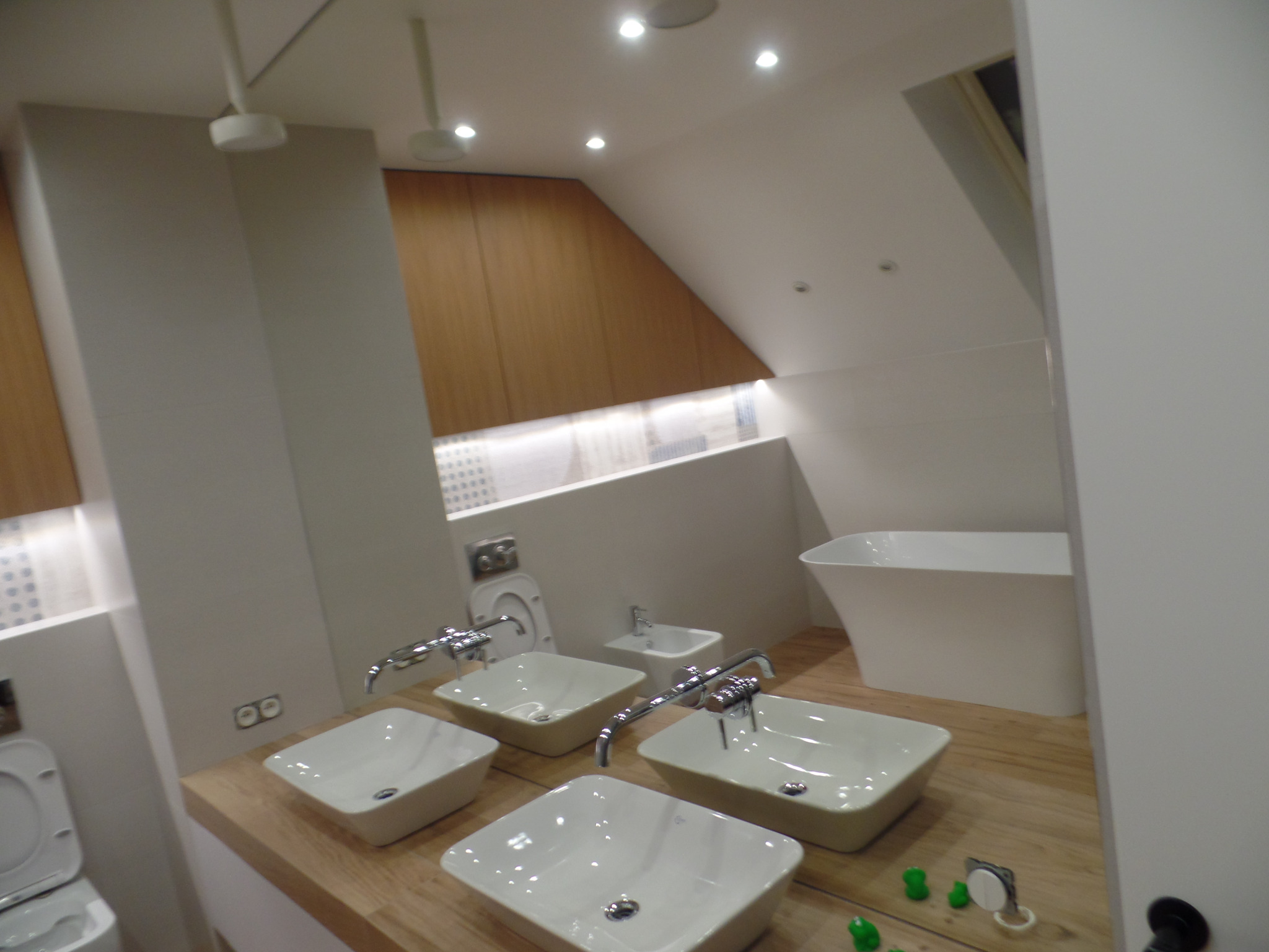 Nowoczesna łazienka w jasnych barwach z czterema umywalkami nablatowymi, toaletą, bidetem i wolnostojącą wanną pod skosem dachu.