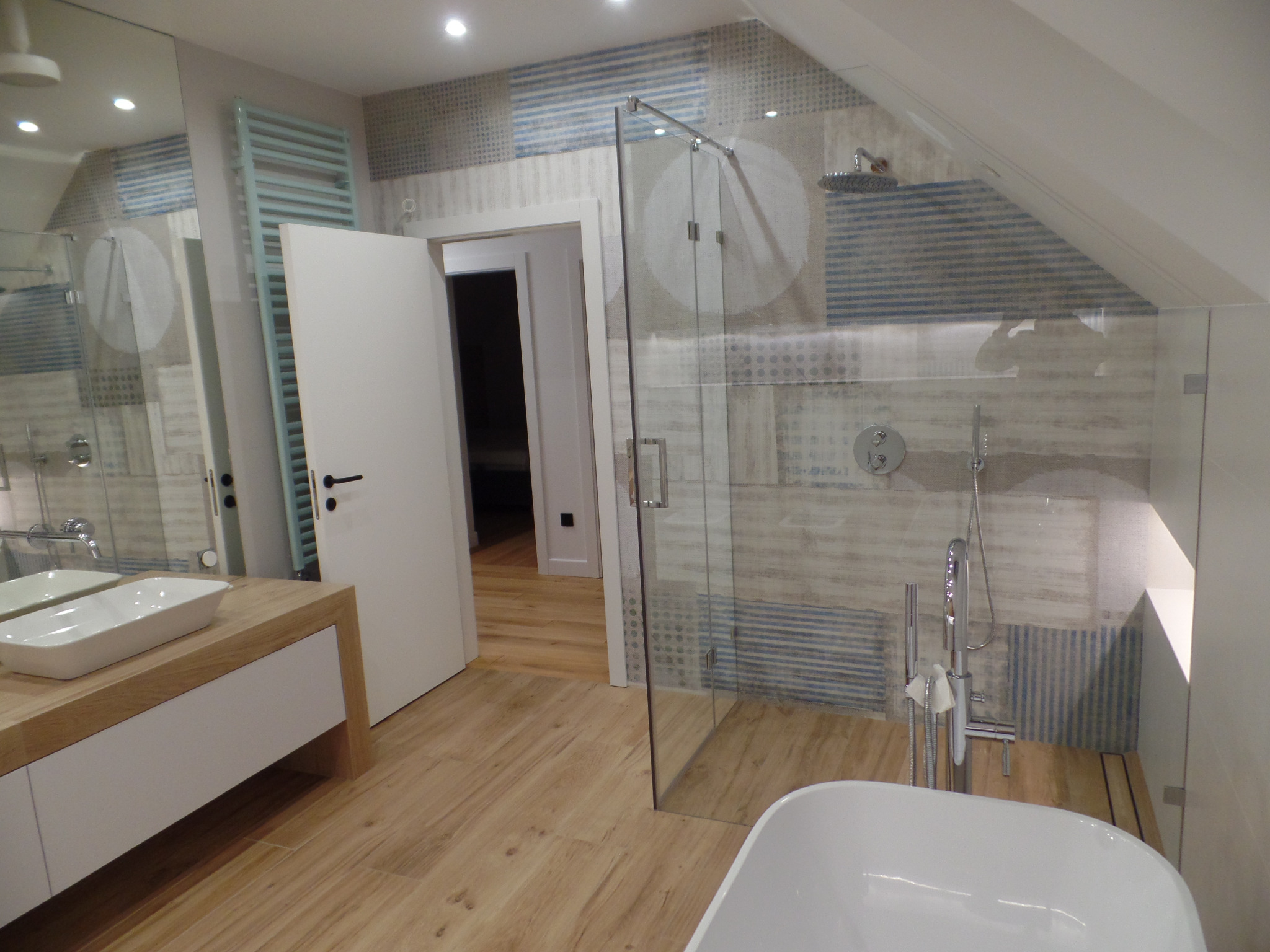 Nowoczesna łazienka z drewnianą podłogą, szklaną kabiną prysznicową i umywalką na szafce z drewnianym blatem. W tle widoczny grzejnik drabinkowy i otwarte drzwi do innego pomieszczenia.