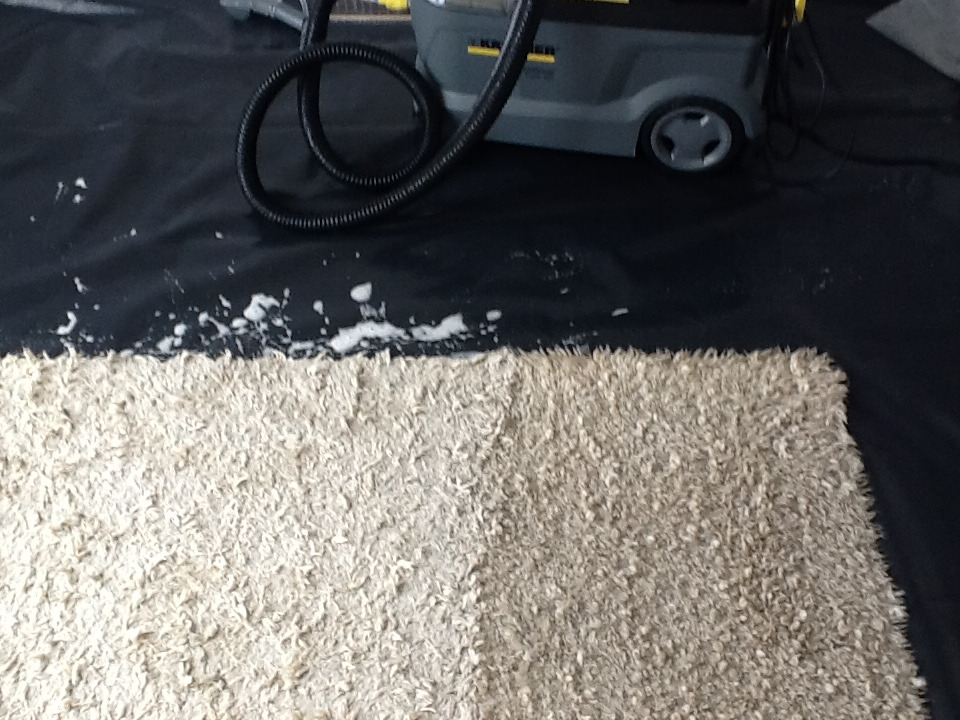 Szary odkurzacz piorący Karcher z wężem na czarnej folii obok fragmentu czyszczonego, beżowego dywanu z widoczną pianą po czyszczeniu.
