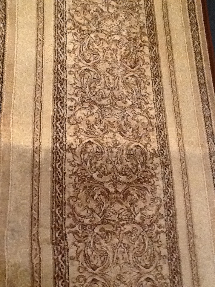Beżowy dywan z symetrycznym, bogatym wzorem w odcieniach brązu, widoczne pionowe pasy po bokach.