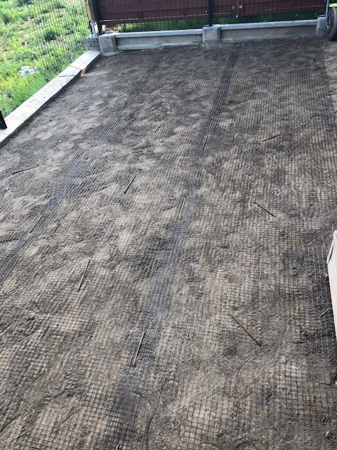 Przygotowana podbudowa pod trawnik z rolki: wyrównany teren z siatką wzmacniającą i wbitymi kołkami mocującymi, widok z góry, na tle ogrodzenia.