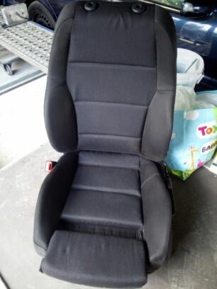 Czarny fotel samochodowy z regulowanym zagłówkiem i widocznym pasem bezpieczeństwa, sfotografowany w warsztatowym otoczeniu z elementami wyposażenia i pudełkiem z zabawkami w tle.