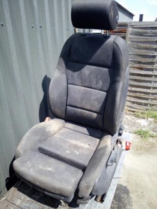 Brudny, używany fotel samochodowy z zagłówkiem, sfotografowany na zewnątrz przy metalowej ścianie.