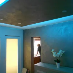 Wykończony sufit z ciemną strukturą i podświetleniem LED, ściany z efektem dekoracyjnym w odcieniach błękitu, białe kwiaty na komodzie, okno z widoczną sylwetką osoby.