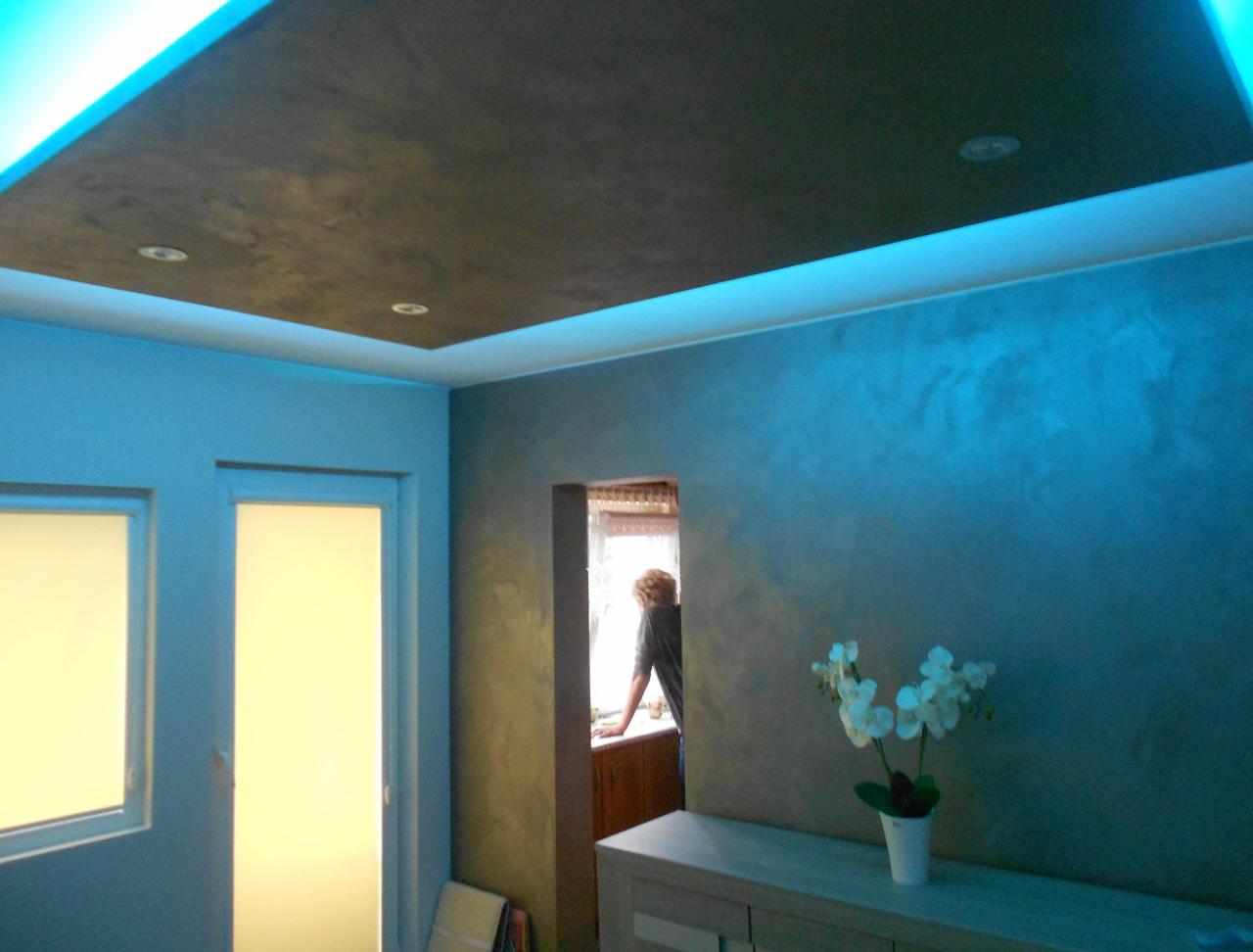 Wykończony sufit z ciemną strukturą i podświetleniem LED, ściany z efektem dekoracyjnym w odcieniach błękitu, białe kwiaty na komodzie, okno z widoczną sylwetką osoby.