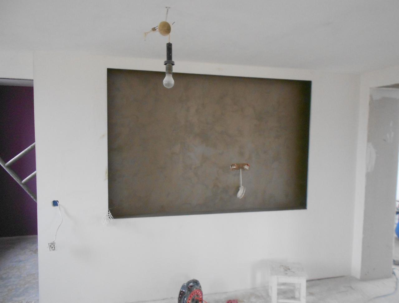 Ściana z wykończoną wnęką w odcieniach brązu, widoczna instalacja elektryczna, surowa żarówka zwisająca z sufitu, w tle widoczny fragment drabiny