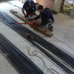 Dwóch pracowników w czerwonych rękawicach tnie pręty zbrojeniowe na drewnianej desce za pomocą specjalnej gilotyny, w tle leżą pęczki prętów oraz szlifierka kątowa, widoczne ślady po usuniętych...