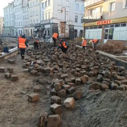 Układanie kostki brukowej na placu miejskim w trakcie renowacji, widoczni pracownicy w pomarańczowych kamizelkach, budynki w tle.