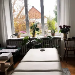 Jasny gabinet z beżowym stołem do masażu, widokiem na ogród przez okno i roślinami doniczkowymi na parapecie.